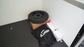 Spare tire resting on a cardboard box in the corner of a cargo trailer, with stacked white boxes and a black bag with a white logo nearby.