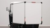 Compact electric delivery vehicle with a tall white enclosed cargo box and black chassis, photographed in a studio setting.
