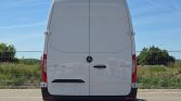 Rear view of a white Mercedes-Benz Sprinter van with dual back doors and high roof, parked on a road with a fence and greenery in the background.
