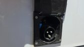 Industrial 3-pin power inlet with protective cover open on a white panel panel edge visible behind it