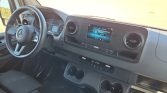 Interior of a Mercedes van: steering wheel, digital dashboard, center touchscreen, round air vents, cupholders at the dash.