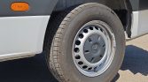 Front-side view of a vehicle wheel with a steel rim, tire mounted on a gray van, showing an orange side marker logo nearby