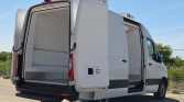 White cargo van with rear double doors wide open, exposing an empty cargo area lined with metal rails and a rubber floor. Side sliding door also open, parked outdoors on a sunny day.