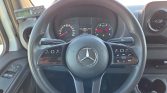Close-up of a Mercedes steering wheel inside a car, with wood trim controls and the instrument cluster in view behind it.