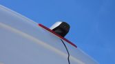 Rear-view camera mounted on the edge of a white vehicle against a clear blue sky, with a visible black cable trailing down.