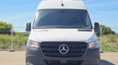 Front view of a white Mercedes-Benz cargo van with a black grille and roof air conditioner, parked outdoors on a sunny day.