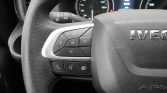 Close-up of a vehicle steering wheel showing a directional pad with OK button and phone/voice control buttons on the left, dashboard gauges in the background (Iveco).
