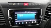 Car infotainment screen showing radio interface (YOU FM) with AM/FM/DAB options, surrounded by control knobs and a red hazard button above.