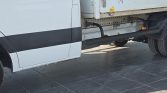 Side view of a white cargo truck showing a black running board under the cab and a metal cargo trailer, parked on dark tiled flooring.