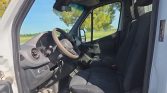 Interior of a white van with the driver’s seat, steering wheel (beige cover), and dashboard visible; rural landscape seen through the open door window.