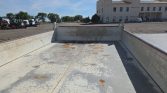 Empty flatbed truck interior with chipped gray paint and rust spots, in an open industrial yard with a beige building in the background.
