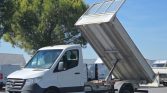 White utility truck with a hydraulically raised dumping bed in a parking lot, blue sky and trees in the background