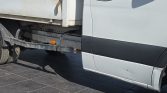 Side view of a white cargo truck showing a black midsection trim, a rusty toolbox on the bed, and exposed undercarriage with dual rear wheels still on the ground on a tiled surface.