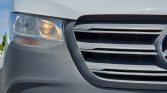 Close-up of a white van's front end, showing the left headlight with its turn signal lit and the grille with a partial Mercedes badge.
