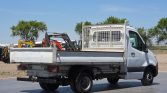 White flatbed truck with a metal mesh divider behind the cab, parked on a lot with construction equipment in the background