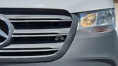 Close-up of a gray car front with grille, emblem, and right headlight visible in daytime light