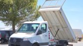White Mercedes-Benz dump truck with its bed raised for unloading, hydraulic piston visible against a blue sky and trees in the background
