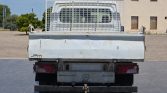 Rear view of a weathered white flatbed truck with a metal cage cargo rack, showing red taillights and a Mercedes-Benz emblem on the bumper in a parking area.