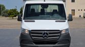 Front view of a white Mercedes cargo van, facing the camera with headlights on, in a paved lot with a beige building in the background.