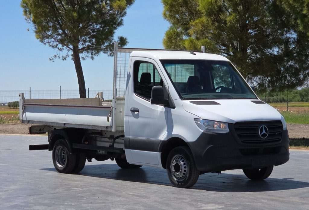 White Mercedes-Benz flatbed truck parked on a concrete lot, with trees and fencing in the background.