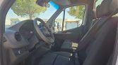 Interior of a van cab with steering wheel, dashboard, and two front seats visible in daylight, outside trees and vehicles seen through the windows.