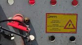Yellow warning plate reads 'Attention Coupe-Circuit' with battery cut-off switch on a gray metal panel, red lever nearby with cables around it.