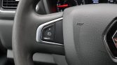 Left side of a car steering wheel with cruise control buttons (+, -, and a speed limiter icon) embedded in a slim silver bezel. The dashboard gauges are blurred in the background, and a horn symbol is visible on the wheel.