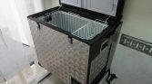 Compact diamond‑plate chest freezer with a black lid, open to reveal white metal wire baskets inside in a utility room.