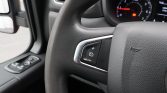 Close-up of a car steering wheel showing cruise control buttons on the left, with the instrument cluster in the background and a red warning light visible.