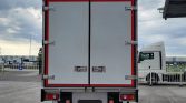 Rear view of a large grey cargo trailer with double doors and red trim, parked in a loading yard beside a white tractor unit.