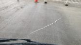 View from inside a car in rain: a diagonal windshield crack with cones and a white fence outside front.