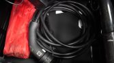 Coiled black EV charging cable with plug in a car trunk beside a red bag