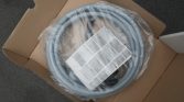 Coiled gray flexible hose wrapped in plastic inside an open cardboard box, with an instruction leaflet resting on top.