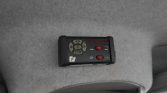 Black handheld remote control with a circular D-pad, two red toggle switches, and a row of red LEDs, mounted on gray car headliner.