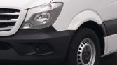 Front left side of a white van showing the headlight, black bumper, and front wheel with a steel rim.