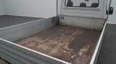 Roll-off truck bed with a weathered wooden floor, gray metal sides, and a mesh cab divider; shows rust and wear along the bed surface.