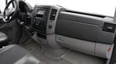 Left-hand drive van interior with steering wheel, gear shifter, and central dashboard featuring radio and climate controls. The gray seats and dusty floor are visible beneath a spacious cabin.
