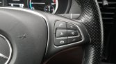 Close-up of a car steering wheel with right-side controls for phone, volume, voice command, and mute buttons on a perforated leather rim (Mercedes)
