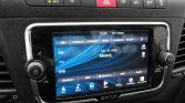 In-dash infotainment touchscreen showing radio interface with blue waveform background and presets above, volume knob on the left.