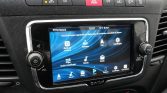 Infotainment touchscreen in a car displaying icons for navigation, media, radio, settings, and backup camera on a blue swirl background, with physical controls and hazard button above.