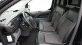 Front interior of a vehicle showing the steering wheel, dashboard, and three front seats with dark upholstery and rubber floor mats start to mid-cabin view from an open door.